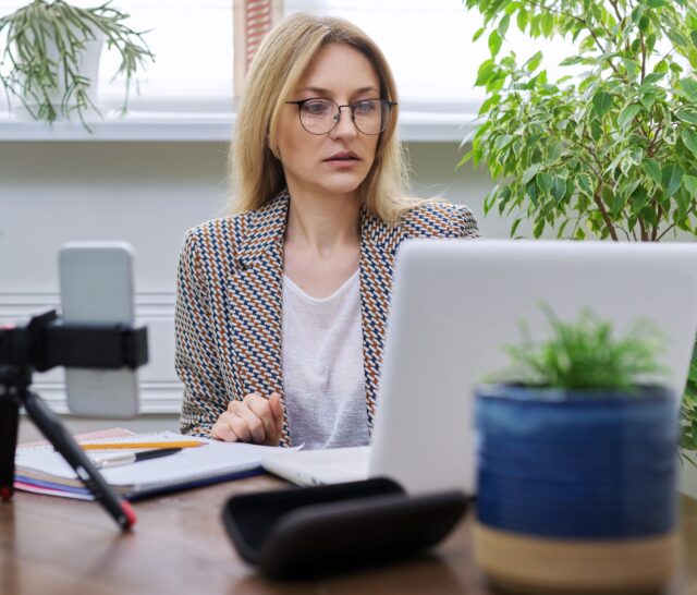 Business woman working at home online using laptop and smartphone. Middle-aged confident female using video call, conference, looking at webcam, business owner, teacher, accountant working remotely